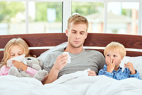 två barn och en pappa sitter bredvid varandra i en säng, alla håller i snytpapper