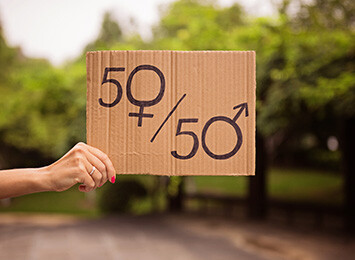 kvinnohand håller en kartongbit där det är skrivet 50/50 där 0:orna är symbolen för kvinna och man, naturen syns i bakgrunden