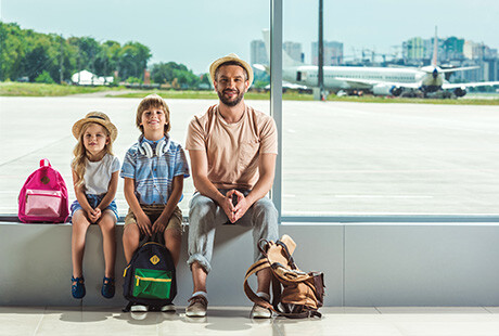 två barn och en pappa sitter inne på en flygplats, utanför syns ett flygplan