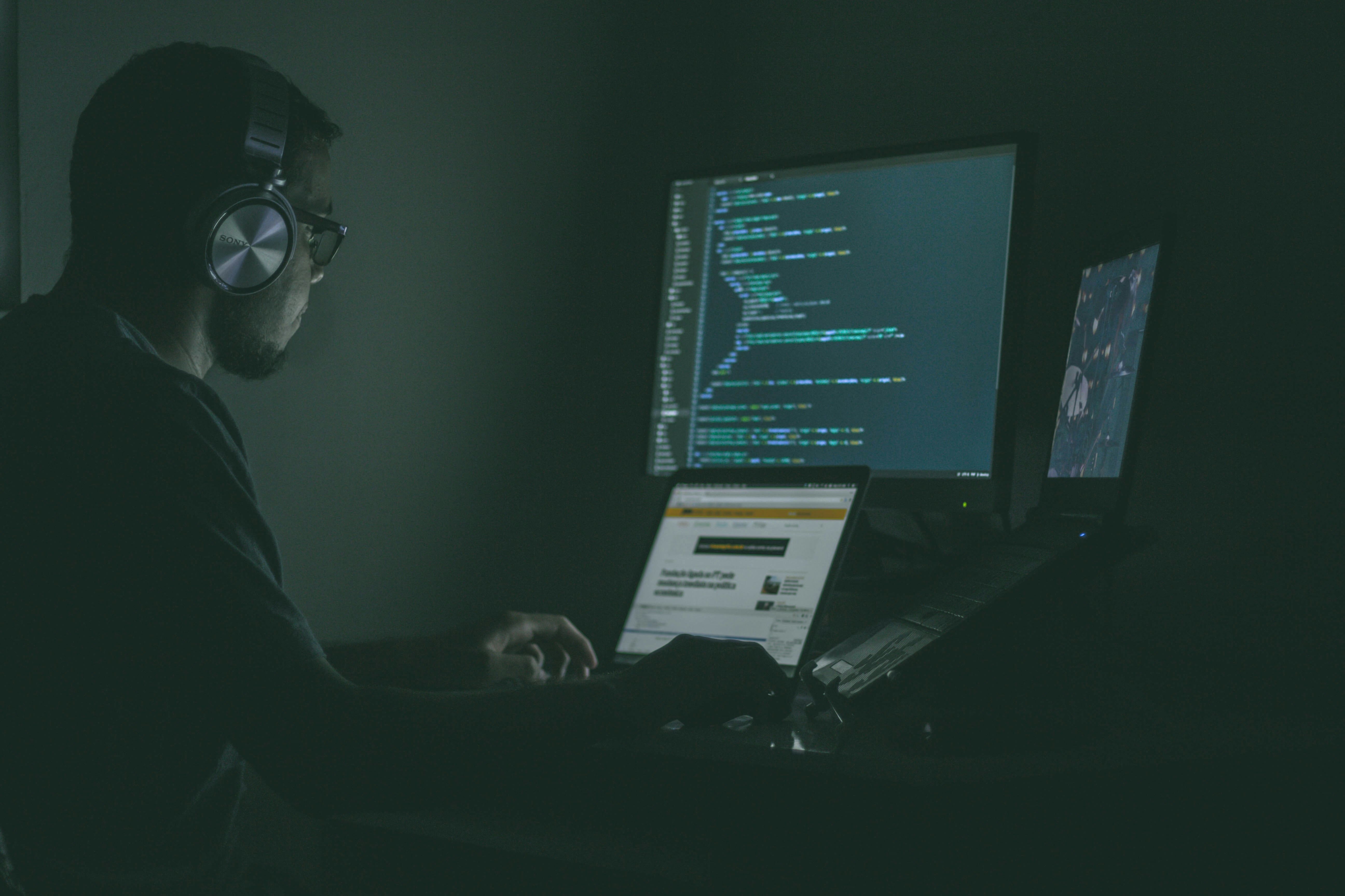 En person i ett mörkt rum som programmerar framför datorskärmar med kod för att säkerställa skydd mot dataintrång.